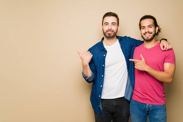 Smiling attractive gay couple in casual clothes pointing next to copy space ad in front of a studio background - Foto, immagini