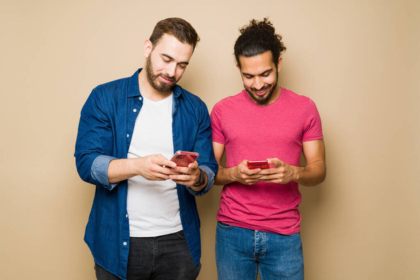 Multiracial gay couple texting on their smartphones and posting on social media against a studio background - Photo, image