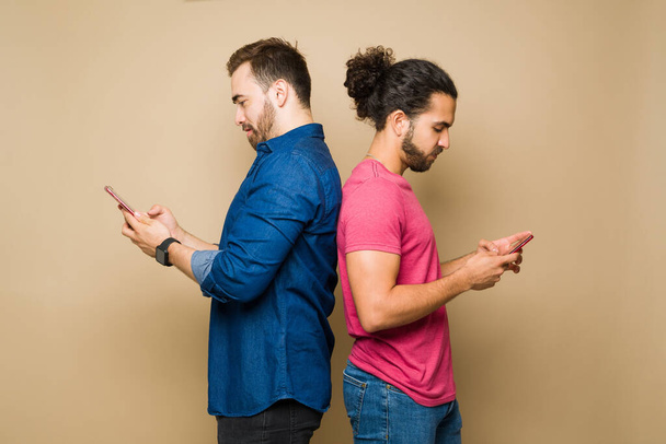 Profile of a caucasian gay man and hispanic boyfriend standing back to back texting on their smartphones - Foto, immagini
