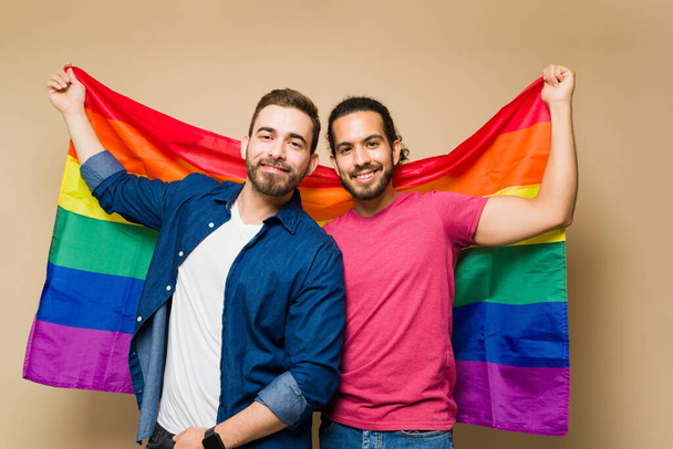 Уверенные геи-мультирасовые мужчины выглядят счастливыми, держа в руках радужный флаг, чтобы поддержать права ЛГБТ перед студийным фоном - Фото, изображение