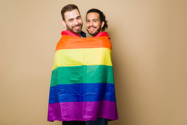 Excited attractive gay couple laughing while playing with a rainbow flag during a LGBT parade - 写真・画像