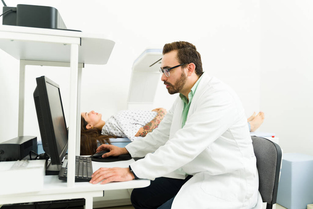 Πλευρική άποψη ενός καυκάσιου αρσενικού ακτινολόγου που κοιτάζει τον υπολογιστή ενώ κάνει ένα τεστ πυκνομετρίας σε μια νεαρή γυναίκα στο εργαστήριο - Φωτογραφία, εικόνα
