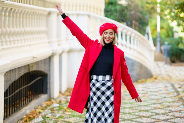 Positive female in red clothes with beret strolling with raised arm and closed eyes on street along white fence in city - Foto, Bild