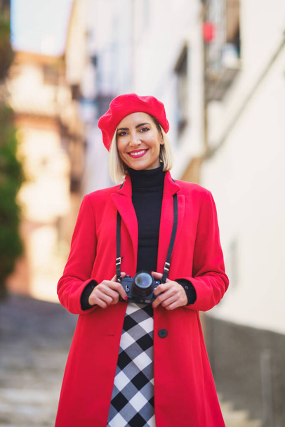Самоуверенная молодая туристка, блондинка в элегантном наряде и берете с фотокамерой, стоящей на узкой улице и улыбающейся в камеру во время экскурсии по старому городу - Фото, изображение