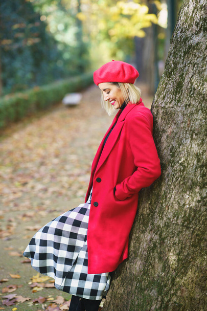 Side view of female standing and hands in pockets of red coat and leaning on tree trunk in autumn park covered with fallen leaves - Fotografie, Obrázek