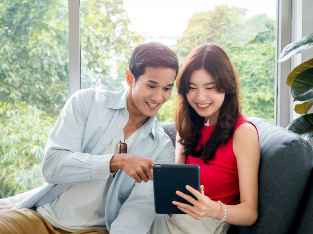 Happy young beautiful Asian woman and handsome man smile together while looking at tablet screen on grey sofa near the glass window at white living room. Couple lover using digital tablet together. - Foto, Imagem
