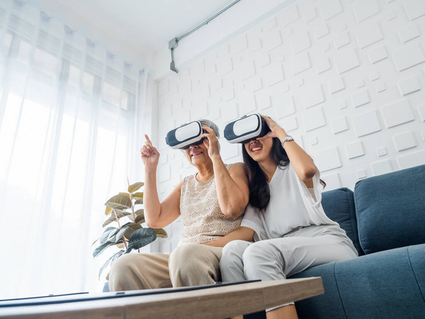 Asian young woman, happy daughter and her mother, old senior female wearing VR glasses, enjoy 3d game online together in white room. Mom and daughter with reality virtual technology lifestyle concept. - Photo, image