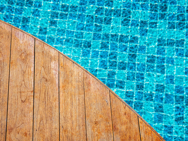 Top view of empty wooden plank table or deck floor curved shape in front of the blurred background of blue mosaic tiles grid pattern in swimming pool. Empty space on poolside, summer background. - Foto, imagen