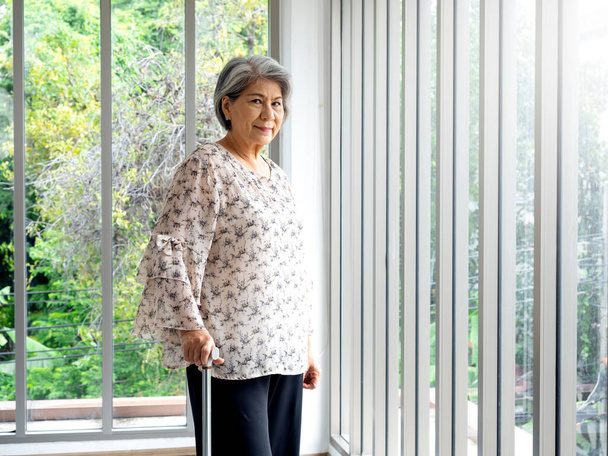Asian senior woman, white hair standing with a cane near the glass window, indoors. Elderly lady patient using walking cane. Strong health, medical care and life insurance concepts. - Photo, image