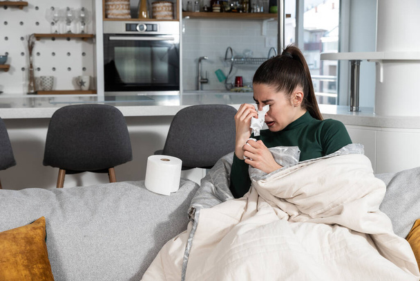 Young sick stressed woman lying at home covered with blanket blowing her nose. Ill female have common cold and flue with runny nose allergy reaction wipe it with paper tissue. - Foto, Bild