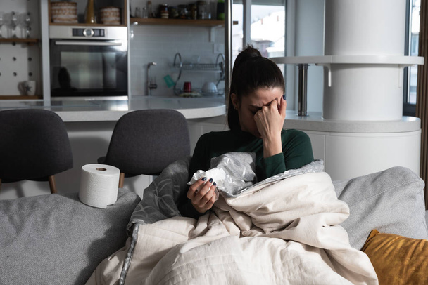 Young sick stressed woman lying at home covered with blanket blowing her nose. Ill female have common cold and flue with runny nose allergy reaction wipe it with paper tissue. - Foto, Imagen