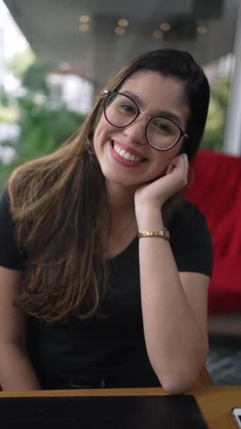 Une jeune femme heureuse souriant à la caméra. Charmante fille des années 20 portant des lunettes assis au café dans la vidéo verticale - Séquence, vidéo