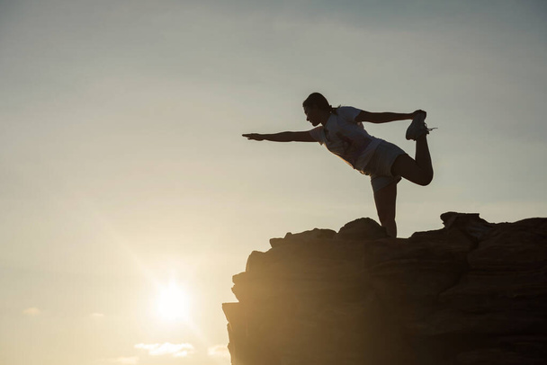 Silhouette athlete sport woman perform yoga exercise on top of hill during sunset after climbing to mountain summit peak. She stretching arms, hands, and legs. Exstreme sport and healthy lifestyle. - Fotografie, Obrázek