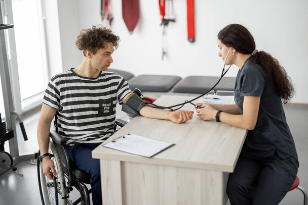 医療従事者は、リハビリテーションセンターで訓練する前に車椅子で若い男の圧力を測定します。医療の概念と負傷者への支援 - 写真・画像