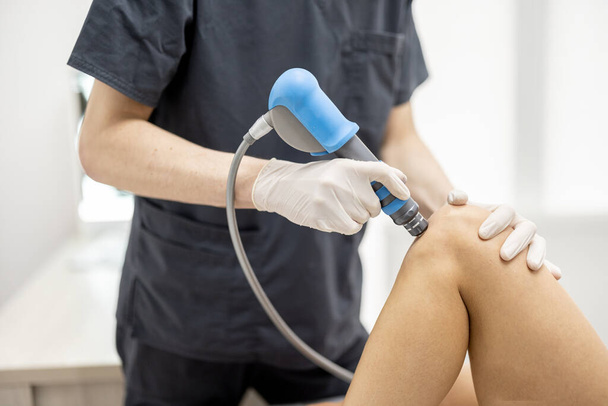 Doctor applies shock wave therapy with special medical equipment on womens knee joints at medical office. Concept of non-invasive technology for treating pain in musculoskeletal system - Photo, Image