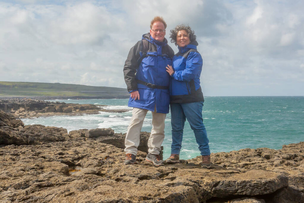 Senior adult couple standing on edge of the rocky cliff against the Atlantic ocean, blue sky with white clouds in blurred background, enjoying a spring day, blue jackets, island of Inis Oirr, Ireland - Foto, Imagem