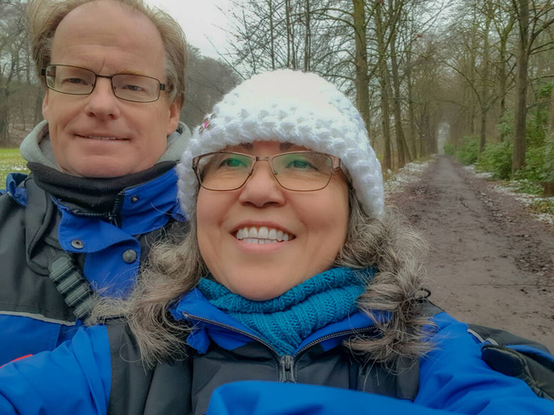 Couple taking a selfie against bare trees with straight path with remnants of snow in background, happy expression, wide smile, winter clothes, crochet hat, blue jackets, wintry day in Netherlands - Photo, Image
