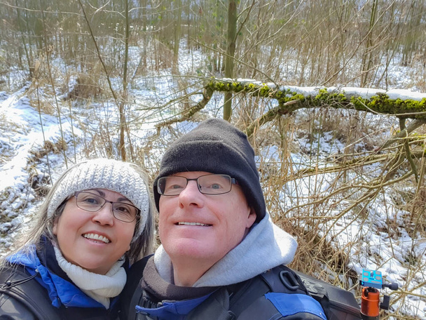 Couple taking a selfie against leafless vegetation and snow on ground, looking up, smiling, wide smile, glasses, winter clothes, crochet headband, black hat, blue jackets, wintry day in Netherlands - Photo, Image
