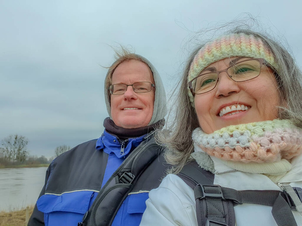 Couple taking a selfie against gray cloudy sky, river in background, looking at camera, smiling, wide smile, glasses, winter clothes, crochet headband and neck, blue jackets, wintry day, Netherlands - Photo, Image