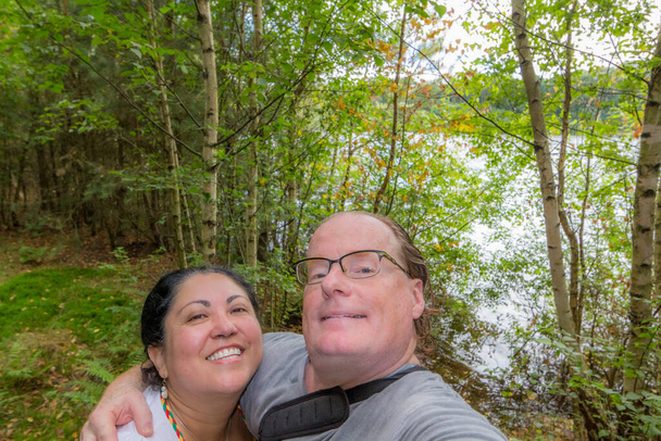 Smiling couple taking a selfie against lush greenery, lake in background, looking up at the camera, wide smile, glasses, casual clothing, sunny summer spring day in nature reserve in Belgium - Photo, image