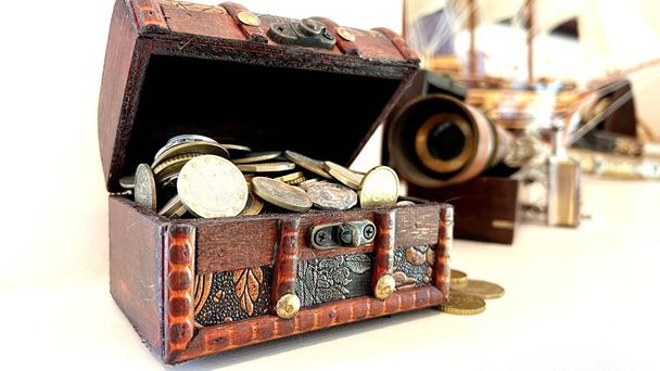 A chest filled with coins, it is ajar from it. Coins scattered in the background, a spyglass is visible, everything is on a white background. Brown wooden box Wealth income earnings saving money. - Photo, Image