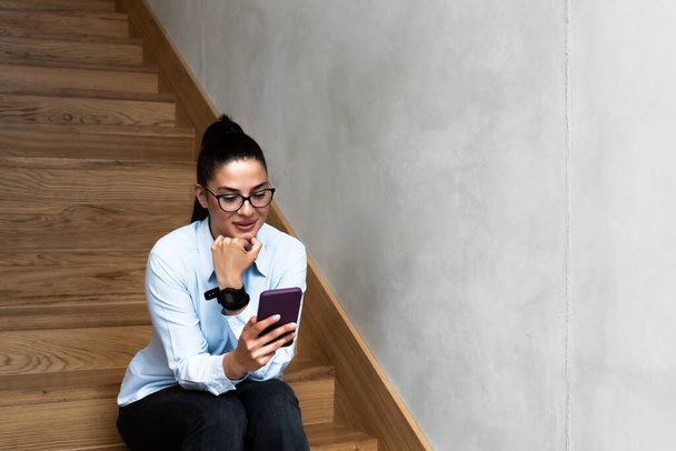 Young expert business woman sitting at the staircase texting and calling her boss to tell that the meeting was well and the deal was signed. Happy female in smart casual sending message on smartphone. - Photo, image