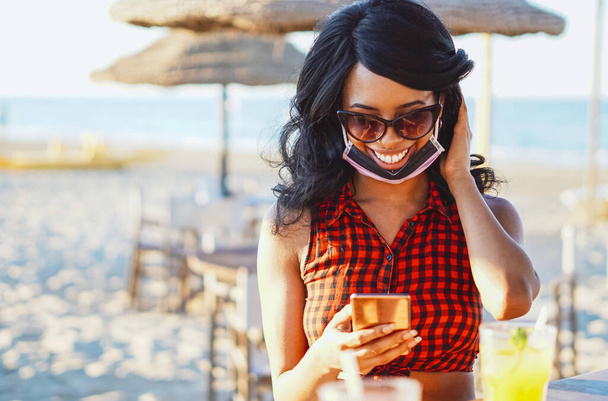 Fille afro-américaine en utilisant smartphone à la plage - Fille assise dans un bar avec masque inférieur sur la similitude et la consommation d'alcool - Photo, image
