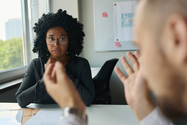 Запутавшаяся молодая многорасовая женщина-менеджер в очках сидит за столом в офисе и общается с коллегой-мужчиной в белой рубашке - Фото, изображение
