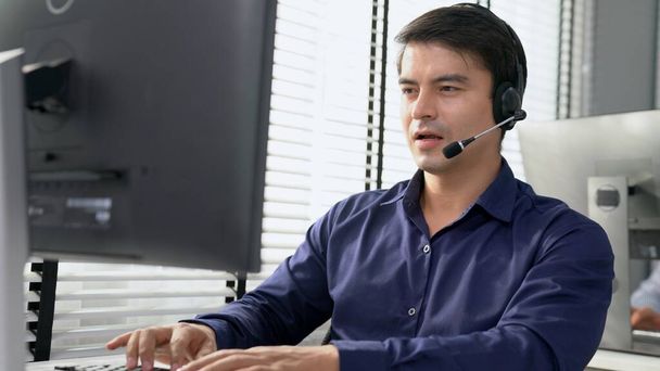 Young competent asian male call center agent working at his computer while simultaneously speaking with customers. Concept of an operator, customer service agent working in the office with headset. - Foto, immagini