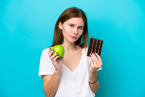 Joven mujer inglesa aislada sobre fondo azul tomando una tableta de chocolate en una mano y una manzana en la otra - Foto, imagen