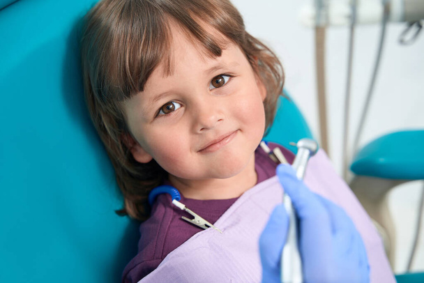 Врач вносит нитриловую перчатку, держа зубоврачебную дрель перед спокойной маленькой девочкой - Фото, изображение