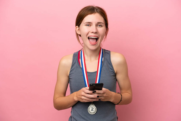 Joven mujer inglesa con medallas aisladas sobre fondo rosa sorprendida y enviando un mensaje - Foto, Imagen