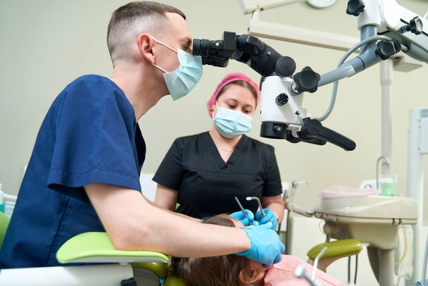 Experienced pedodontist examining child teeth with dental microscope in presence of female assistant - Zdjęcie, obraz