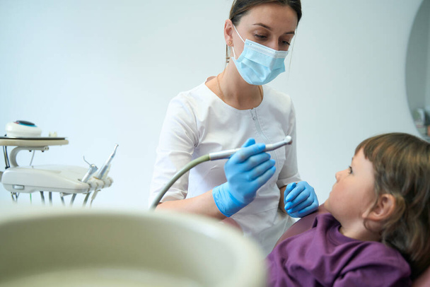 Pediatric dentist in nitrile gloves holding drill handpiece and staring at little patient in chair - Zdjęcie, obraz