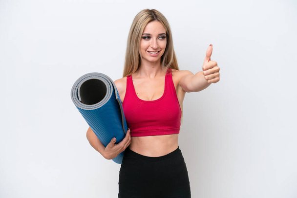 Young sport blonde woman going to yoga classes while holding a mat isolated on white background giving a thumbs up gesture - Photo, Image