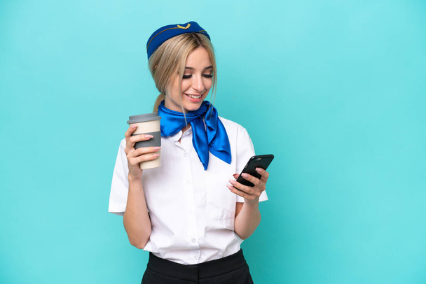 Airplane blonde stewardess woman isolated on blue background holding coffee to take away and a mobile - Photo, image
