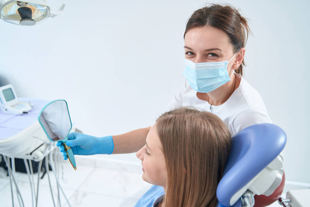 Professional stomatologist holding hand mirror in front of adolescent girl seated in dental chair - Foto, Imagem