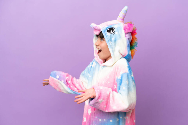 Little kid wearing a unicorn pajama isolated on purple background with surprise facial expression - 写真・画像