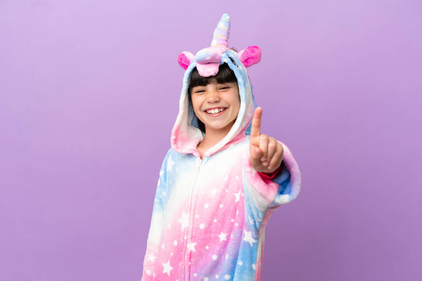 Egyszarvú pizsamát viselő, lila alapon izolált kisgyerek, aki az ujját mutogatja és emeli. - Fotó, kép