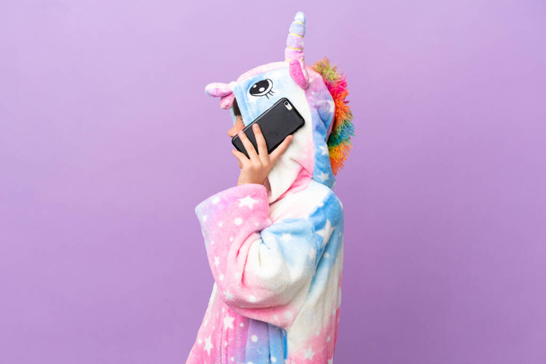 Little kid wearing a unicorn pajama isolated on purple background keeping a conversation with the mobile phone with someone - Photo, Image