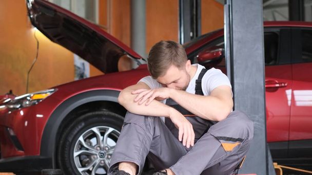 Jeune homme européen épuisé mécanicien automobile. Le mécanicien se repose au travail. Service, atelier de réparation. Vidéo 4k - Photo, image