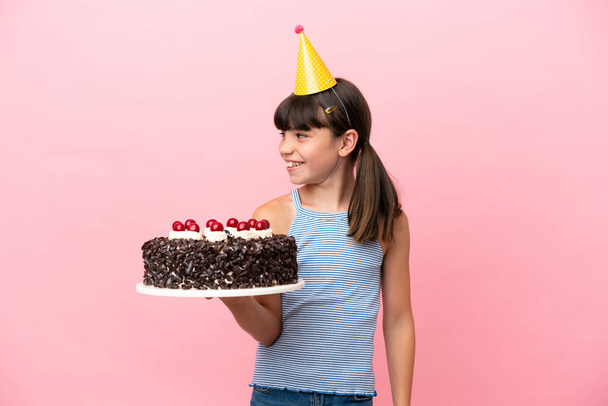 Klein blank kind met verjaardagstaart geïsoleerd in roze achtergrond kijken naar de zijkant en glimlachen - Foto, afbeelding