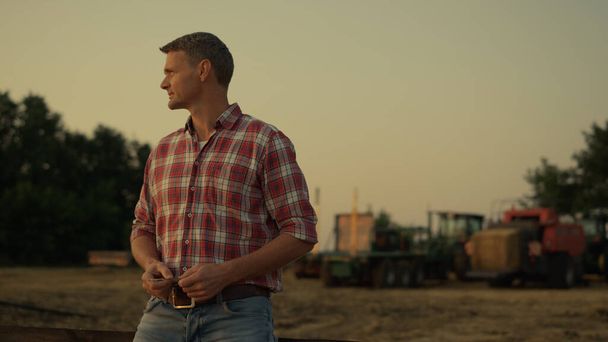 Farmář hledající pšeničné pole při zlatém letním západu slunce. Úspěšný agrární podnikatel stojí na zemědělské půdě u průmyslových strojů a kontroluje pěstované obiloviny. Moderní agrobyznys koncept - Fotografie, Obrázek
