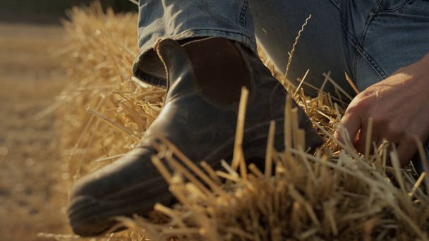 Closeup farmer dusty boot on hay stack closeup. Field worker resting on dry bale after hard work. Unrecognized man agriculture professional relaxing inspecting stubble farmland. Male leg on straw. - Foto, imagen
