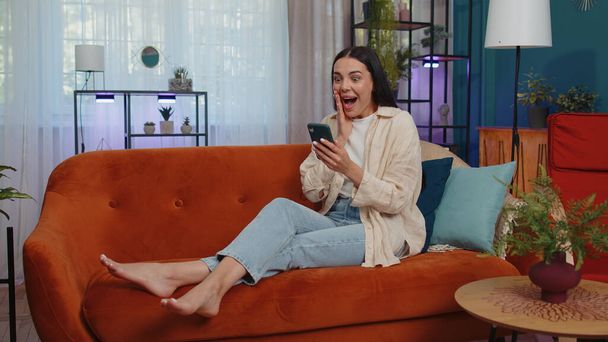 Oh my God Wow. Excited happy joyful winner girl use smartphone typing browsing found out great win good news celebrate victory. Young woman with mobile phone lying at home in room on orange couch - Valokuva, kuva