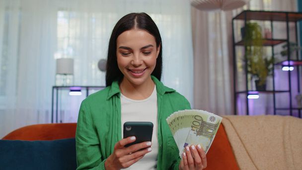 Planning family budget. Smiling caucasian girl counting money euro cash, use smartphone calculate domestic bills at home. Joyful young woman satisfied of income saves money for planned vacation, gifts - Foto, afbeelding