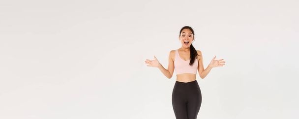 Full length of surprised and amazed female athelte, asian fitness girl in sportsbra and leggings looking wondered, smiling excited and spread hands sideways, find out about gym special discounts. - Photo, Image