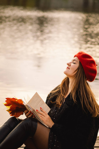 Женщина в красном берете читает книгу о деревянном понтоне. Осенний сезон. Женщина с модной одеждой отдыхает у воды, читает книгу и пьет кофе. Осеннее настроение. Осенняя концепция. Осенний день. - Фото, изображение