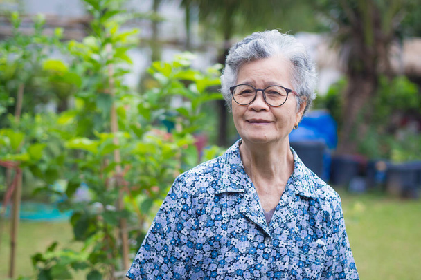 Старшая женщина в очках с короткими седыми волосами улыбается и смотрит в камеру, стоя в саду. Пространство для текста. Концепция пожилых людей и здравоохранения. - Фото, изображение