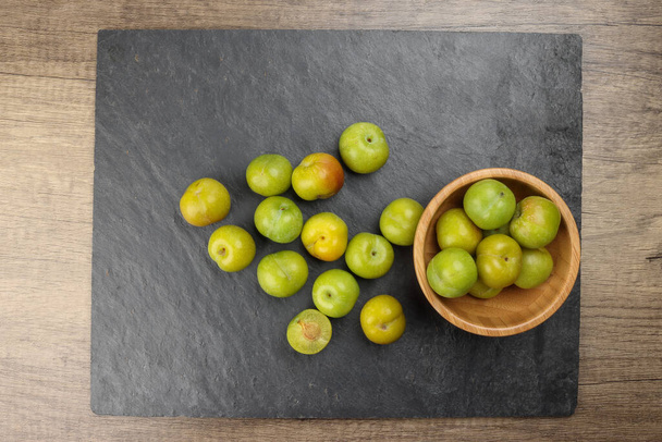 Small mini green fresh juicy plum fruit in wooden bowl on black slate stone board wooden table cut slice half  - Foto, afbeelding